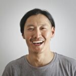 A joyful Asian man laughing against a neutral gray backdrop.